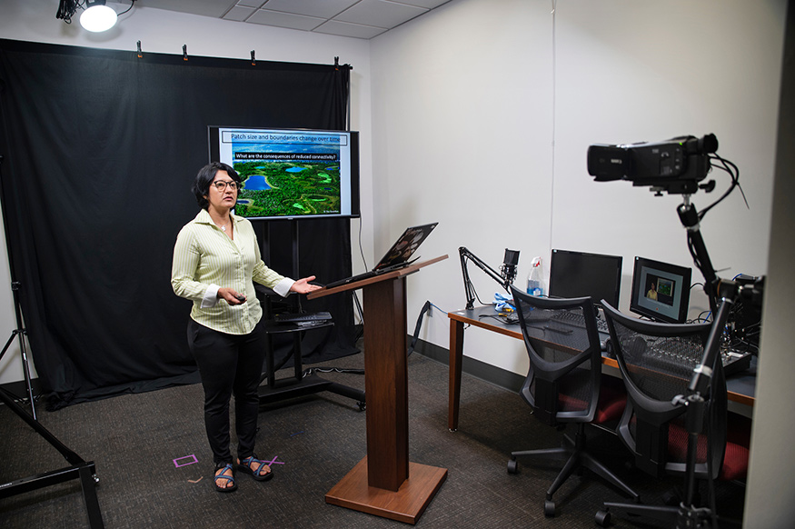 A woman stands in front of a video camera with television monitors in the background as she records a talk.